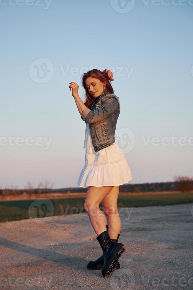 rödhårig kvinna i kjol stående och Framställ för en kamera på de väg på kväll tid foto