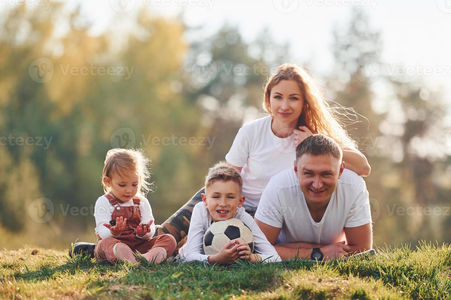 med fotboll boll. Lycklig familj liggande ner utomhus nära de skog. med dotter och son foto