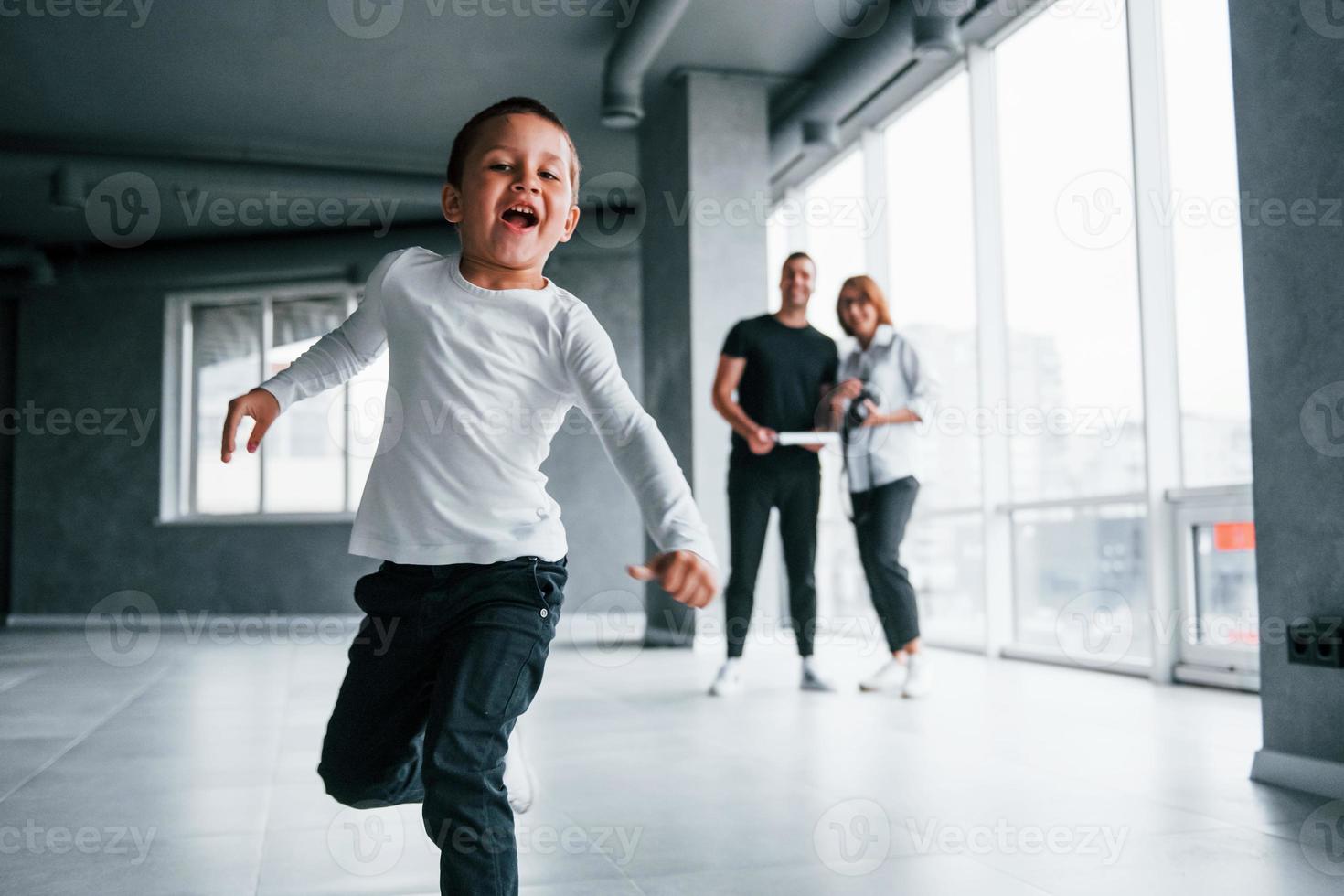 kvinna i formell kläder och med kamera i hand stående inuti av tömma rum med man och liten pojke den där löpning och har roligt foto