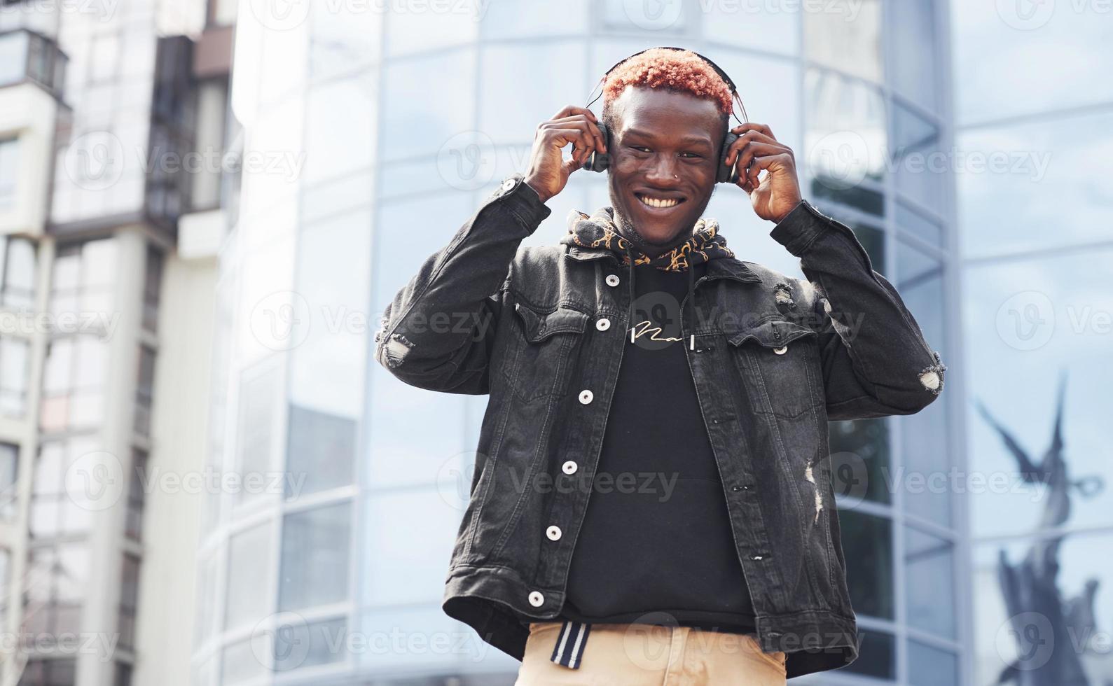 lyssnande till musik i hörlurar. ung afrikansk amerikan man i svart jacka utomhus i de stad stående mot modern företag byggnad foto