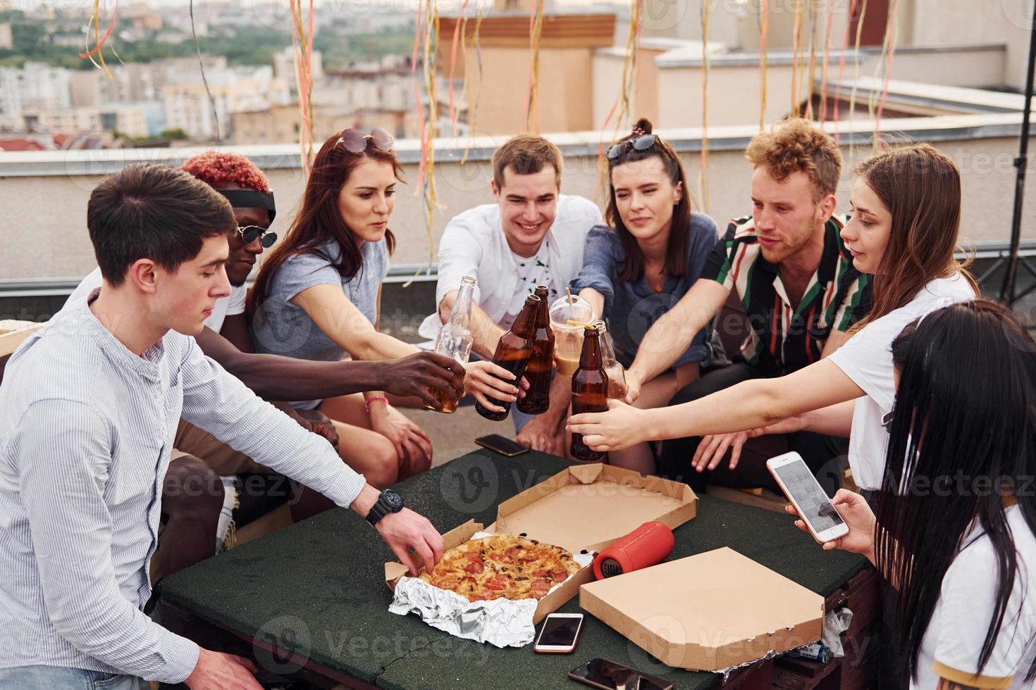 håller på med Skål förbi flaskor med öl. grupp av ung människor i tillfällig kläder ha en fest på taket tillsammans på dagtid foto