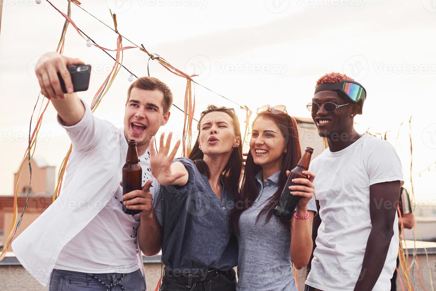 stående tillsammans och tar selfie. grupp av ung människor i tillfällig kläder ha en fest på taket tillsammans på dagtid foto