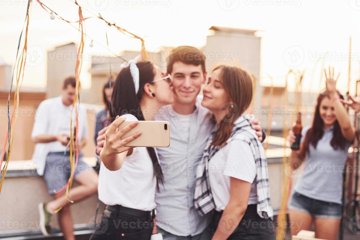 två flickor kissing ett man. tar selfie. grupp av ung människor i tillfällig kläder ha en fest på taket tillsammans på dagtid foto