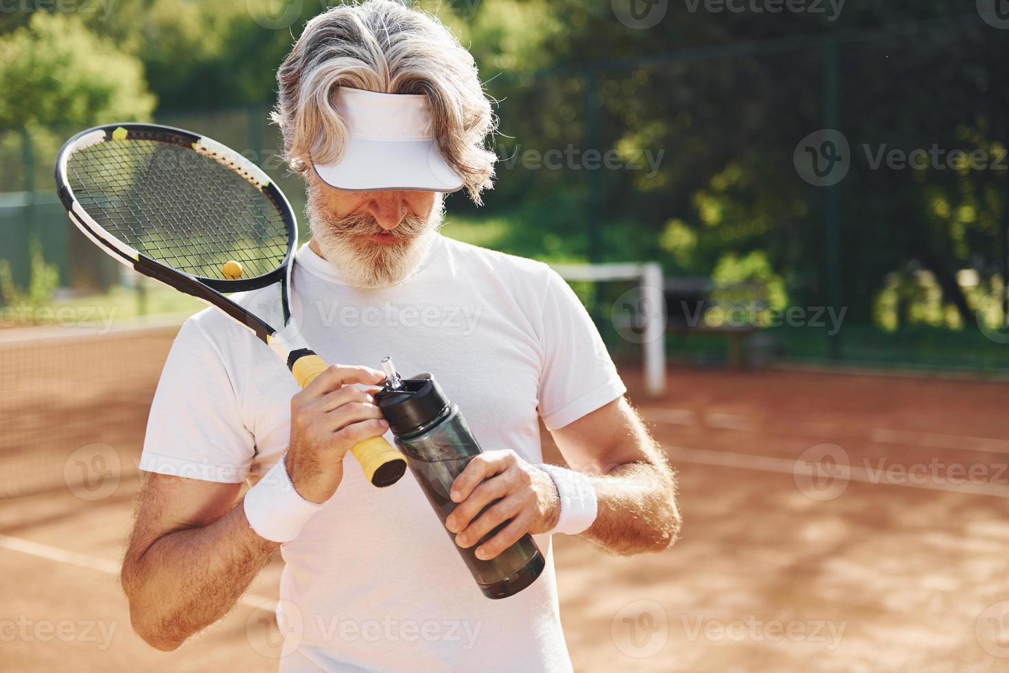 tar en ha sönder och dricka vatten. senior modern eleganta man med racket utomhus på tennis domstol på dagtid foto