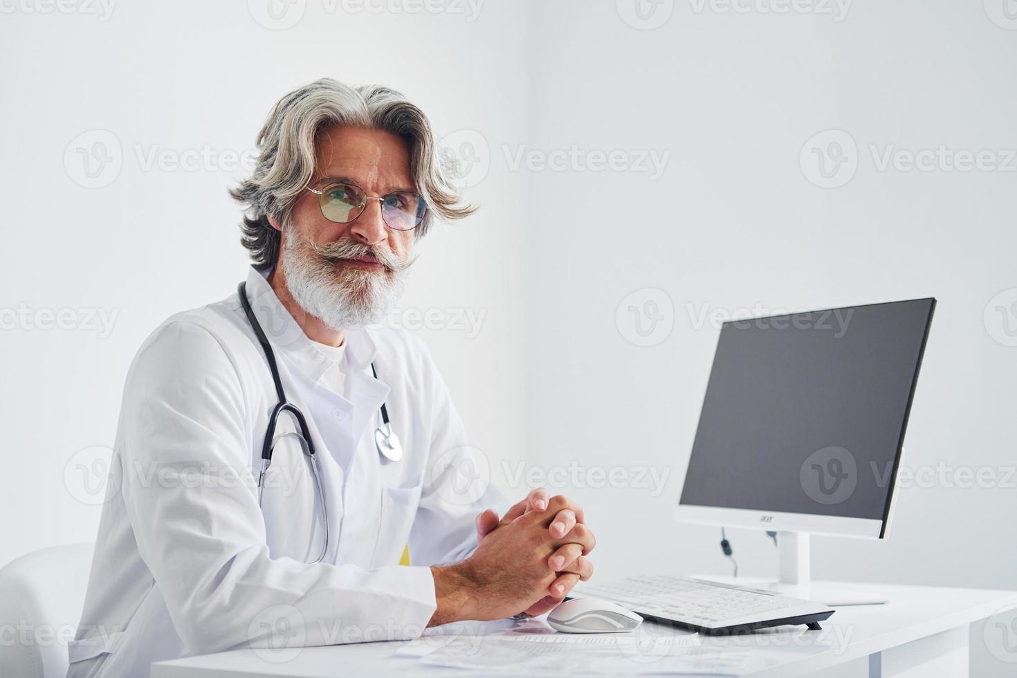porträtt av senior manlig läkare med grå hår och skägg i vit täcka är inomhus i klinik foto
