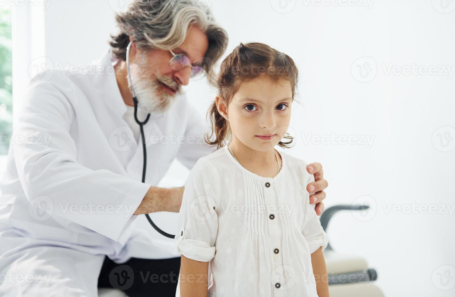 senior manlig läkare med grå hår och skägg i vit täcka mätning hjärta Betygsätta av liten flicka förbi stetoskop inomhus i klinik foto