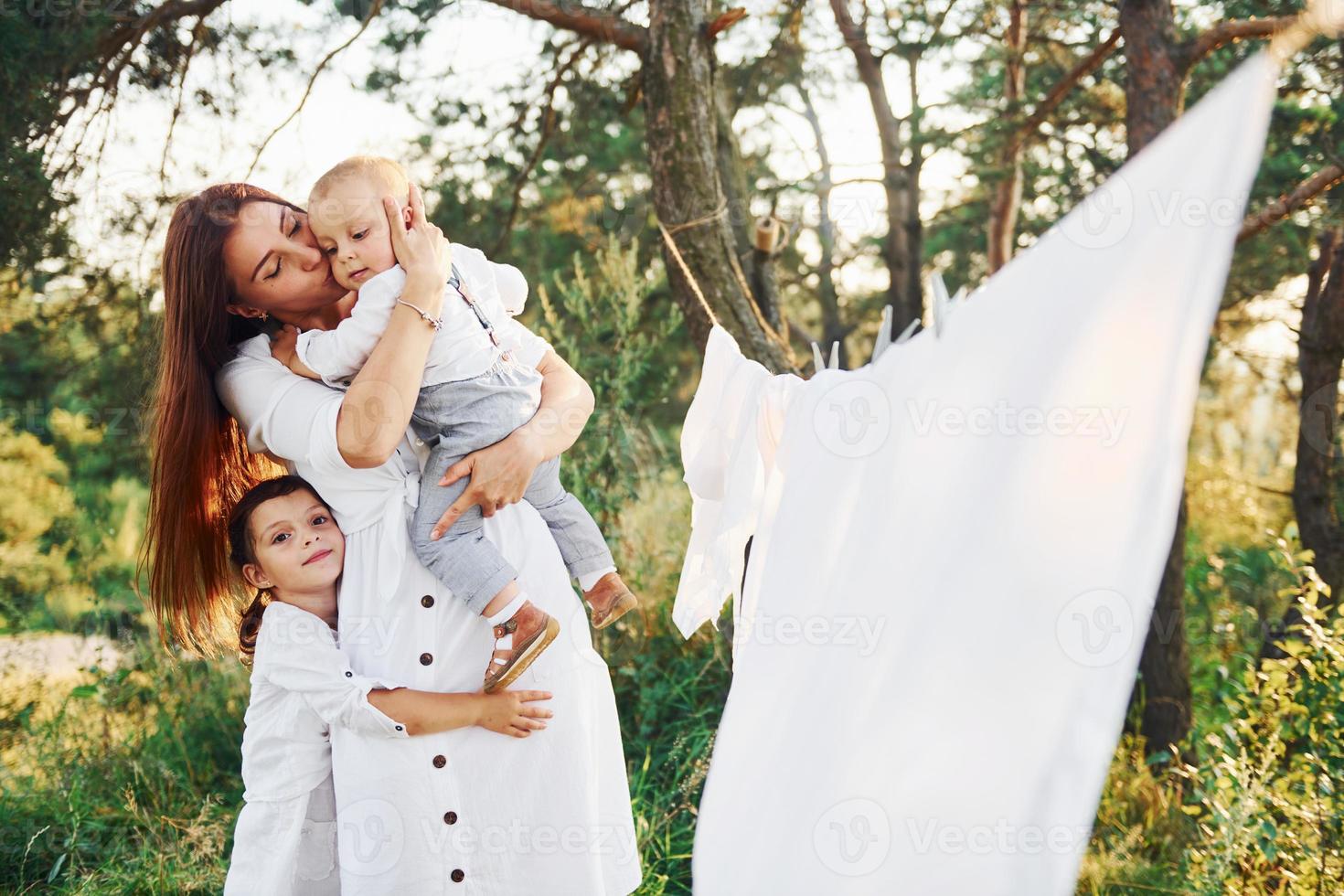 hemmafru torkning kläder. ung mor med henne liten dotter och son är utomhus i de skog. skön solsken foto