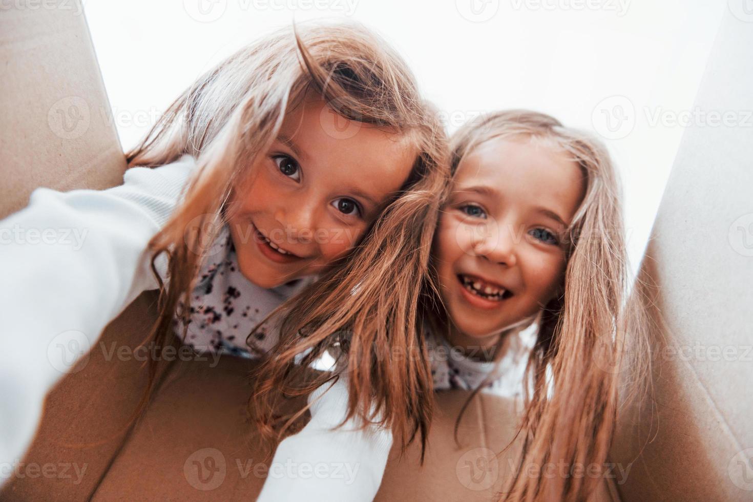 ser inuti av papper låda. två söt liten flickor inomhus på Hem tillsammans. barn har roligt foto