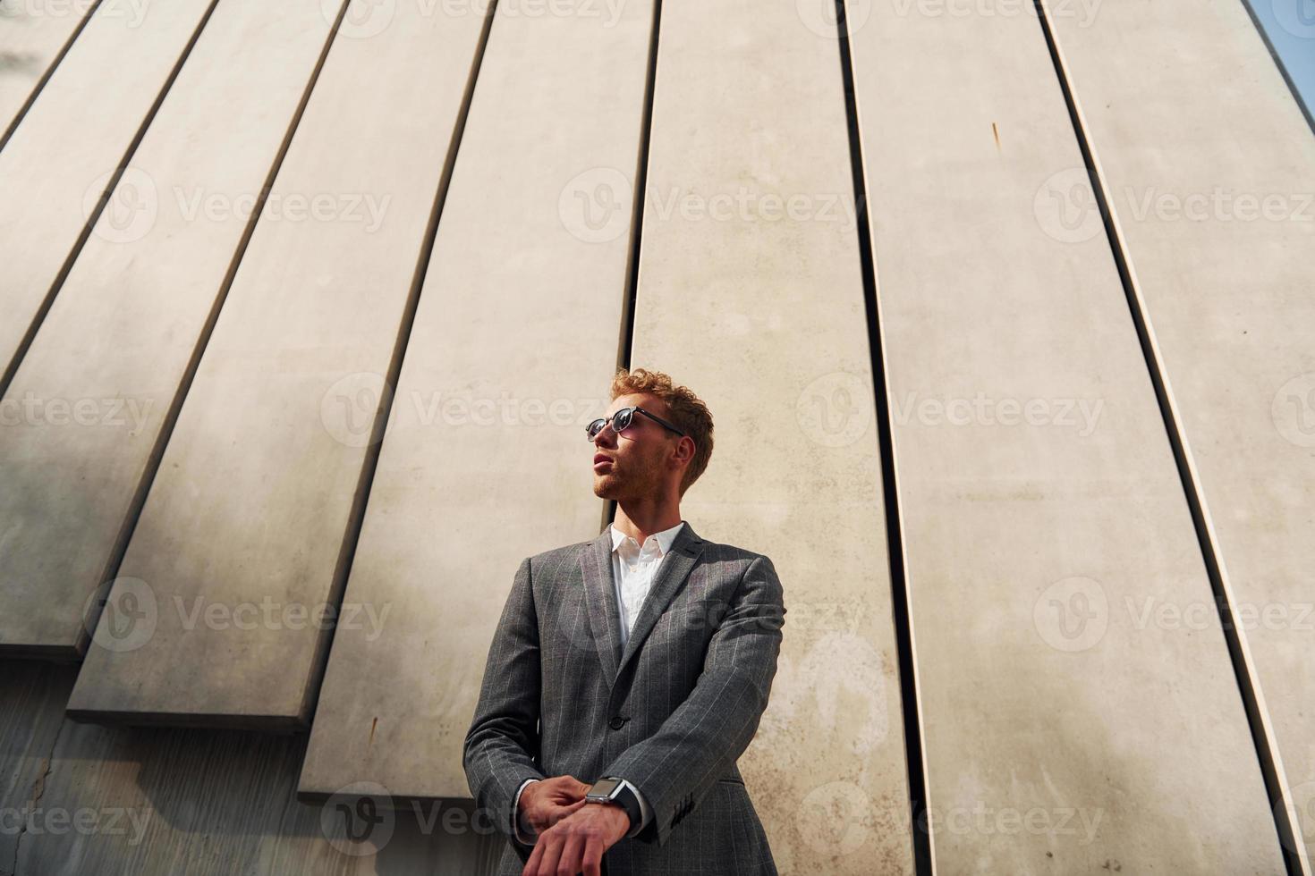 stående mot byggnad vägg. ung affärsman i grå formell ha på sig är utomhus i de stad foto