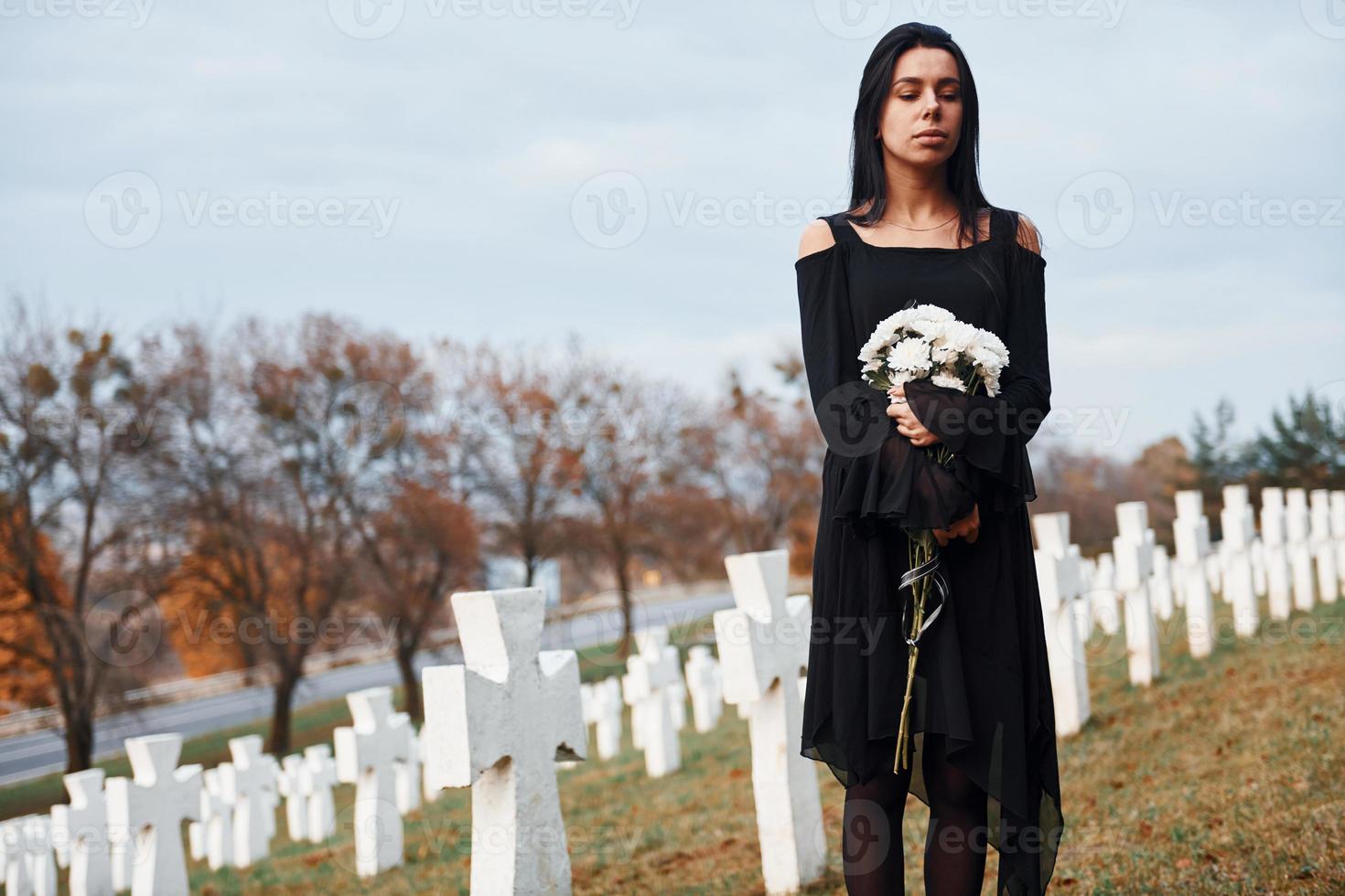 med blommor i händer. ung kvinna i svart kläder besöker kyrkogård med många vit går över. uppfattning av begravning och död foto