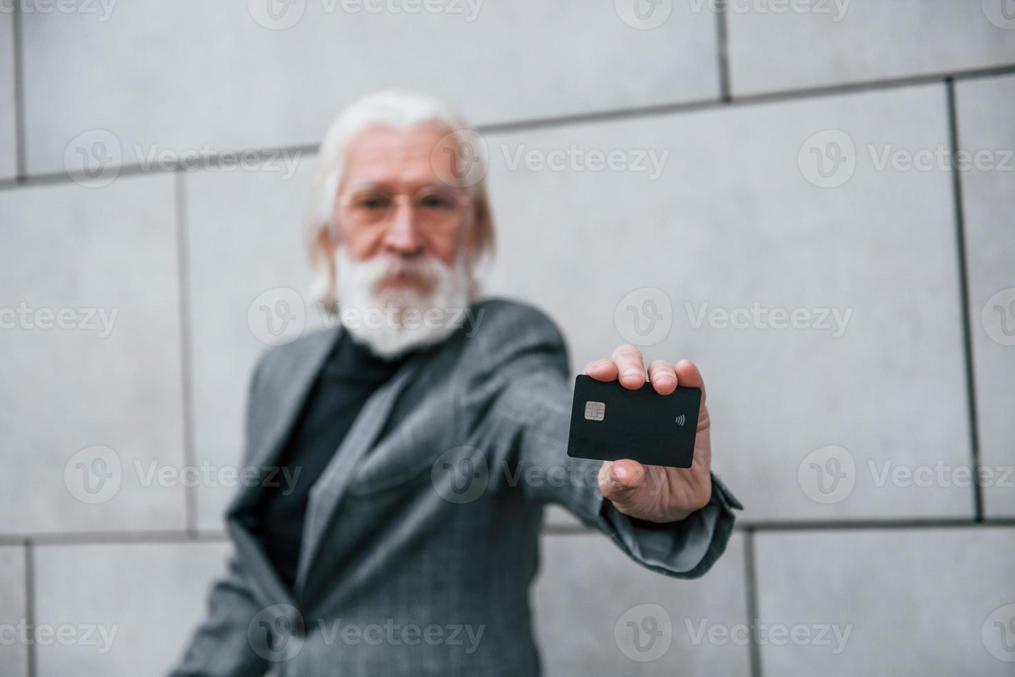 senior affärsman i formell kläder, med grå hår och skägg är utomhus innehar kreditera kort foto