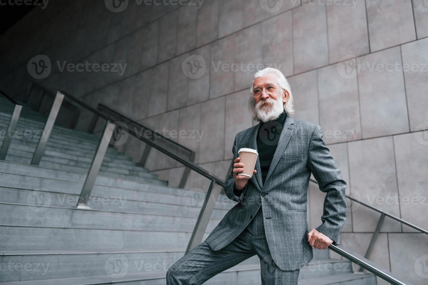 senior affärsman i formell kläder, med grå hår och skägg är utomhus och innehar kopp av dryck foto