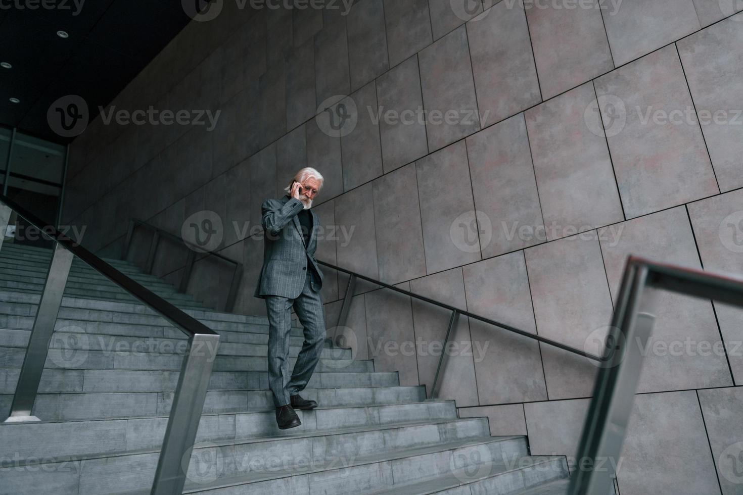 senior affärsman i formell kläder, med grå hår och skägg är utomhus samtal förbi telefon foto