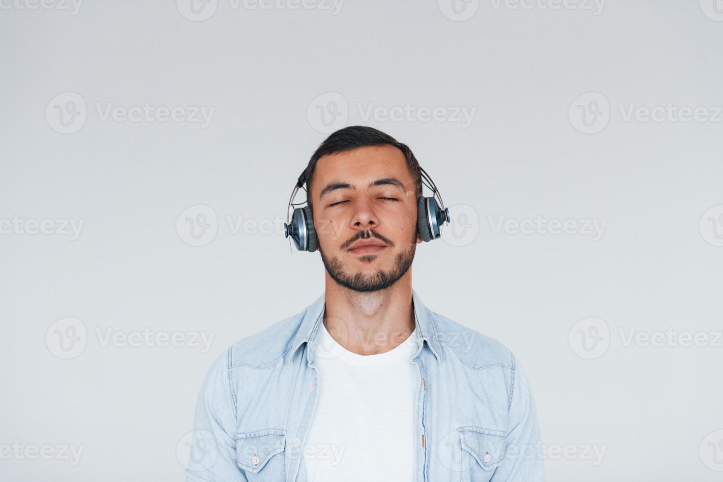 lyssnar till de musik i hörlurar. ung stilig man stående inomhus mot vit bakgrund foto