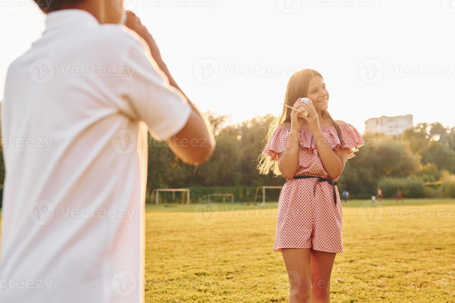 pojke med flicka har roligt förbi använder sig av tenn kan telefon medan stående utomhus på de fält foto