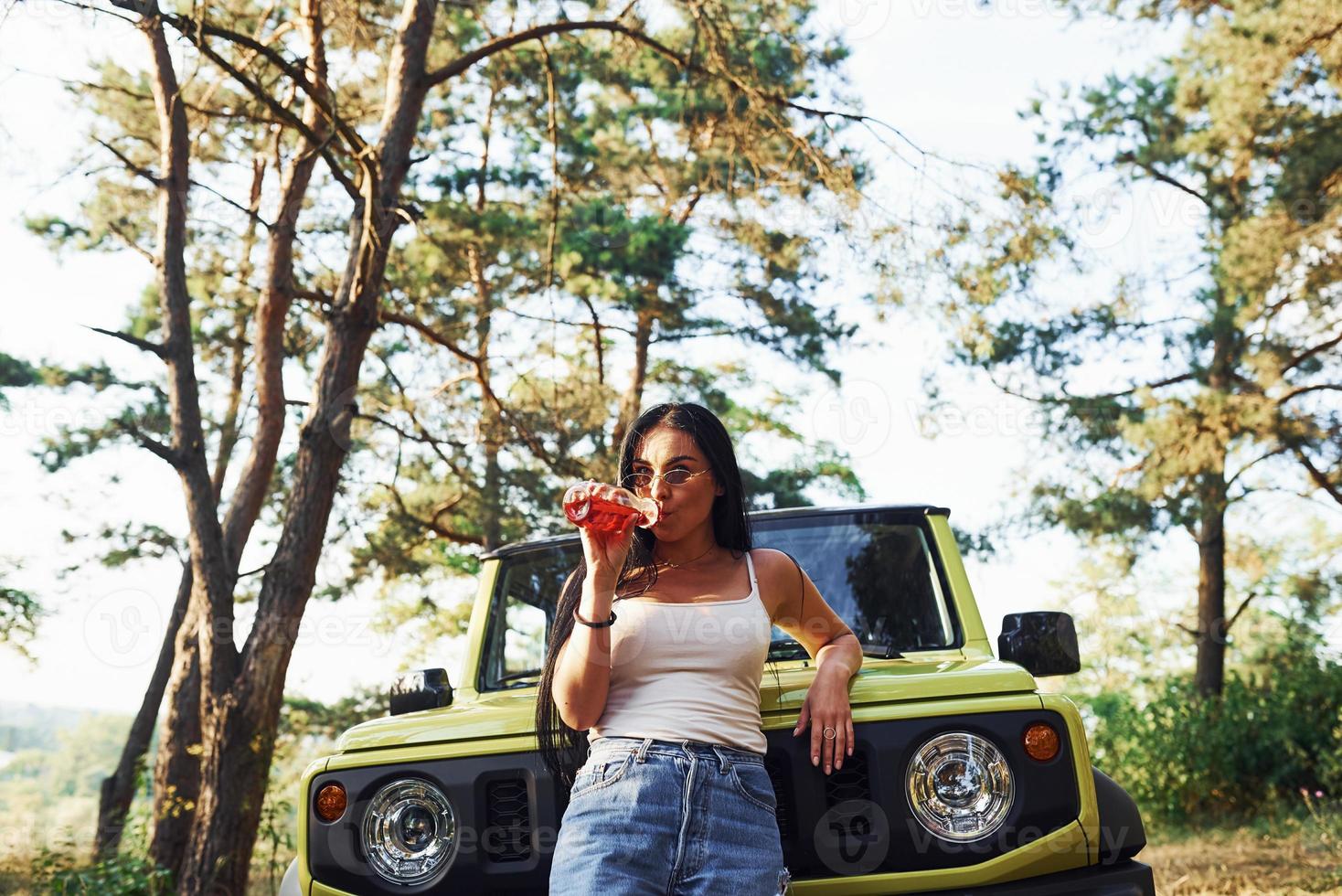 blond med alkohol i hand ha en promenad i de skog mot grön jeep foto