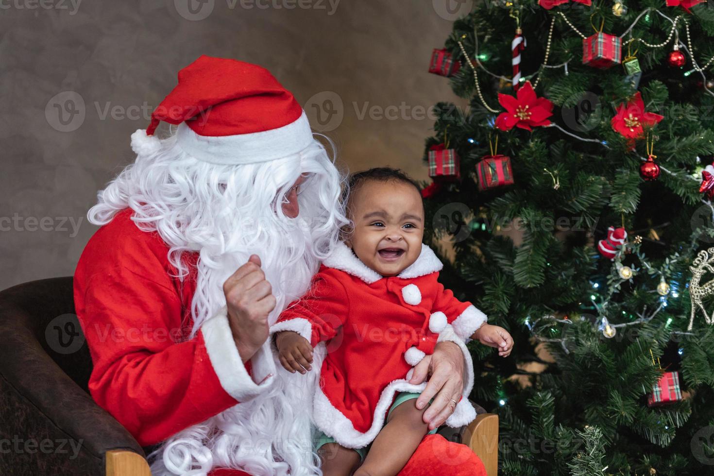 Jultomten lyfter upp den glada lilla flickan och skrattar glatt med fullt dekorerad julgran på baksidan för säsongsfirande koncept foto