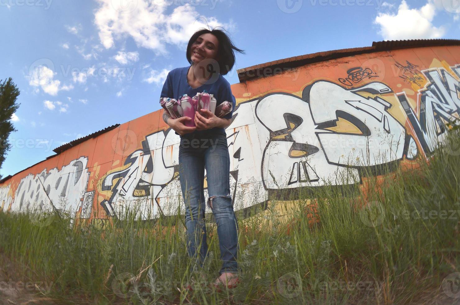 porträtt av ett emotionell ung flicka med svart hår och piercingar. en vid vinkel Foto av en flicka med aerosol måla burkar i de händer på en graffiti vägg bakgrund. en modern porträtt av en fisköga lins
