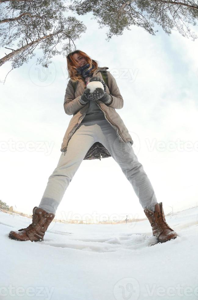 en ung och glad caucasian flicka i en brun täcka innehar en snöboll i främre av en horisont linje mellan de himmel och en frysta sjö i vinter. fisköga Foto