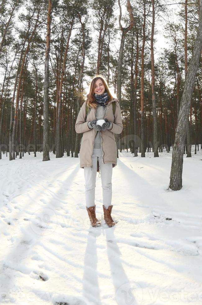 en ung och glad caucasian flicka i en brun täcka innehar en snöboll i en snötäckt skog i vinter. fisköga Foto
