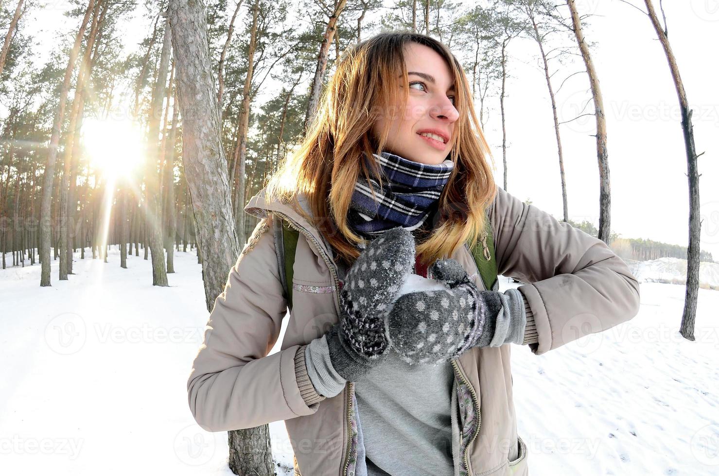 en ung och glad caucasian flicka i en brun täcka skulpterar en snöboll i en snötäckt skog i vinter. spel med snö i de öppen luft. fisköga Foto