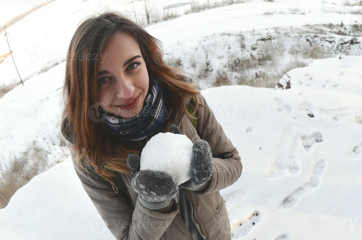 en ung och glad caucasian flicka i en brun täcka innehar en snöboll i främre av en horisont linje mellan de himmel och en frysta sjö i vinter. fisköga Foto