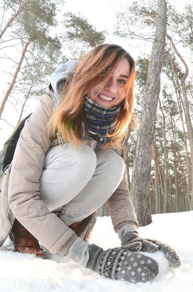 en ung och glad caucasian flicka i en brun täcka skulpterar en snöboll i en snötäckt skog i vinter. spel med snö i de öppen luft. fisköga Foto