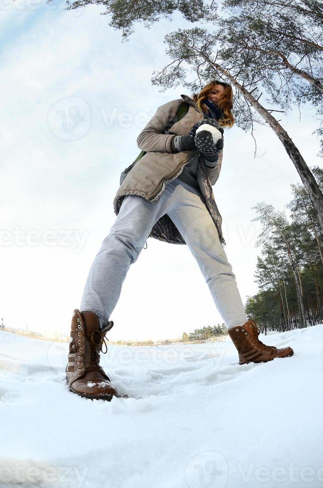 en ung och glad caucasian flicka i en brun täcka innehar en snöboll i främre av en horisont linje mellan de himmel och en frysta sjö i vinter. fisköga Foto