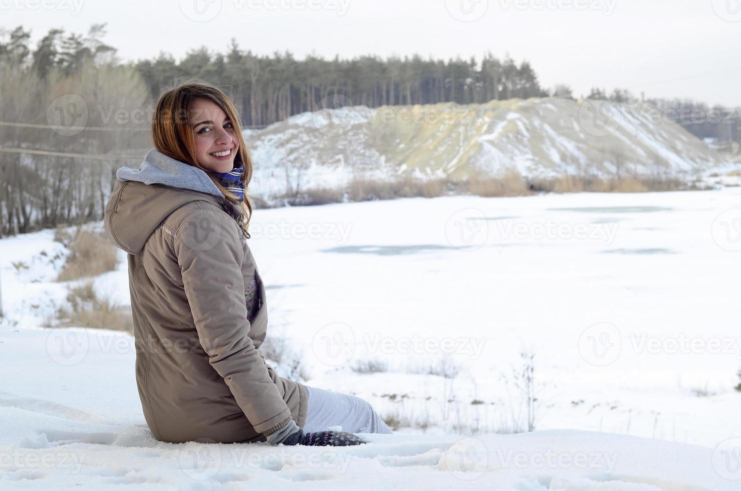 en ung caucasian flicka i en brun täcka är Sammanträde nära en klippa i de bakgrund av en horisont linje mellan de himmel och en frysta sjö i vinter- tid foto