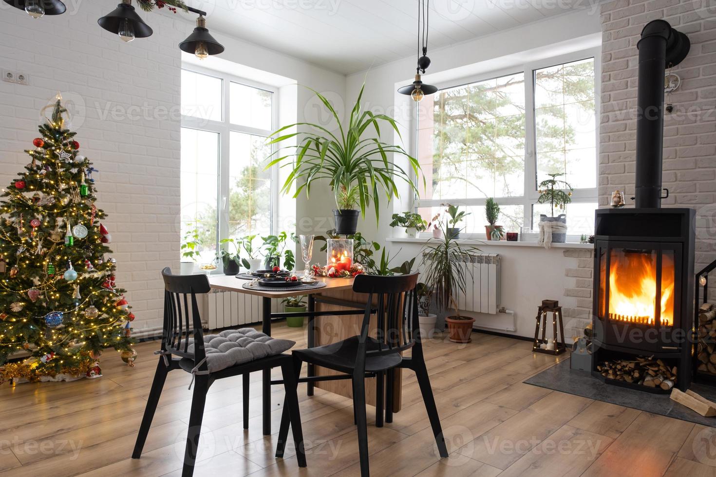 festlig interiör av hus är dekorerad för jul och ny år i loft stil med svart spis, öppen spis, jul träd. värma studio rum med uppsättning tabell, brinnande trä, mysigt och uppvärmning av Hem foto