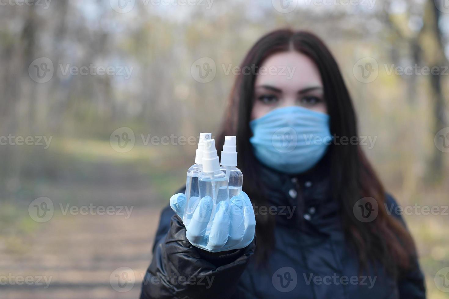 ung kvinna i skyddande mask visar desinfektionsmedel spray flaskor utomhus i vår trä foto