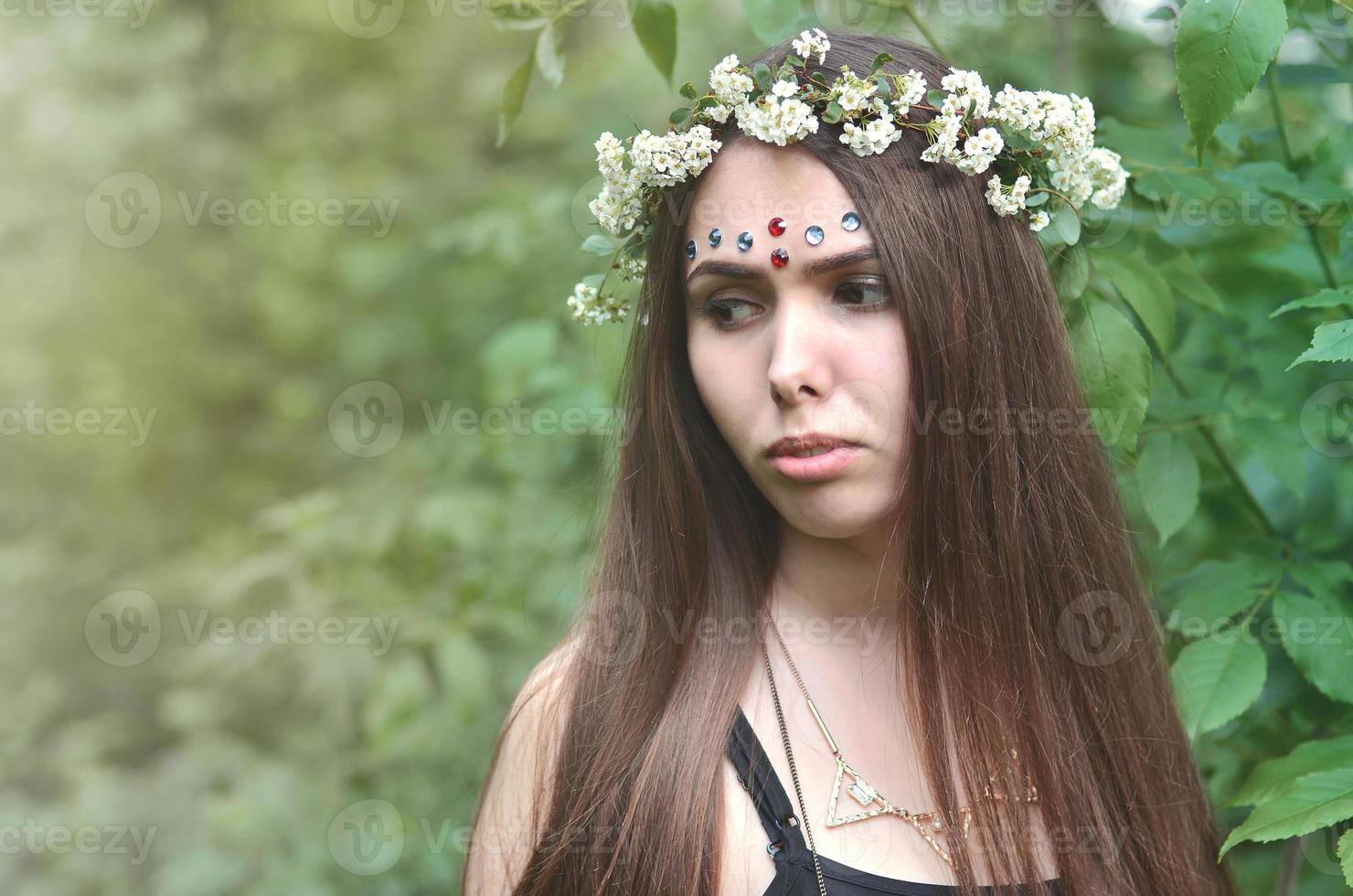 en skog bild av en skön ung brunett av europeisk utseende med mörk brun ögon och stor mun. på de flickans huvud är bär en blommig krans, på henne panna skinande dekorationer foto