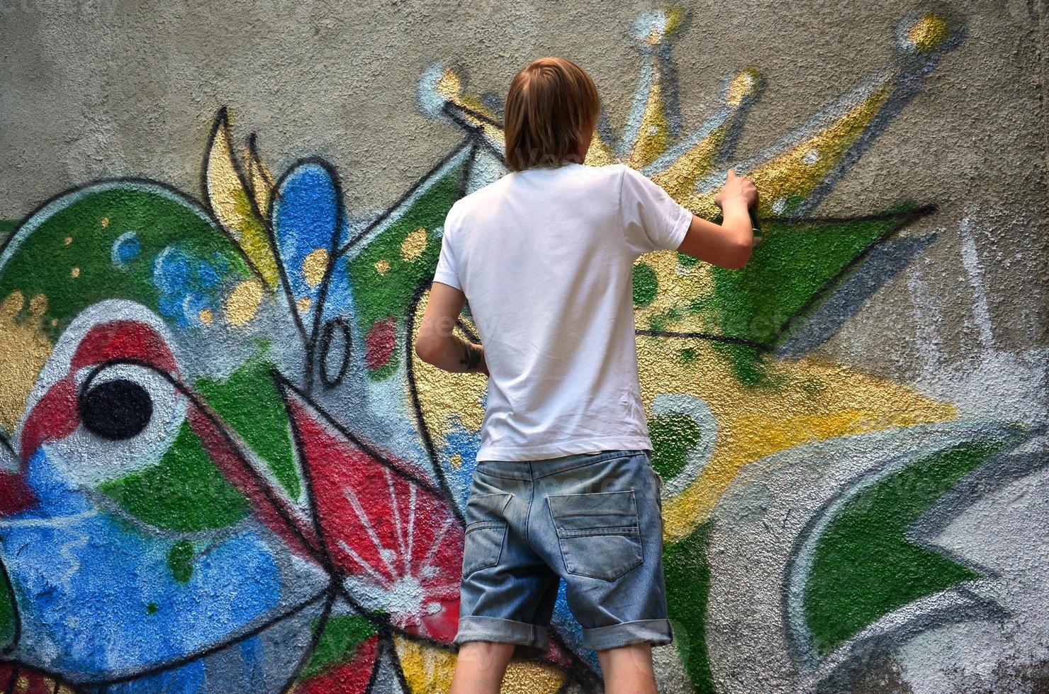 Foto av en ung kille i denim shorts och en vit skjorta. de kille drar på de graffiti vägg en teckning med aerosol målarfärger av olika färger. de begrepp av huliganism och skada till fast egendom