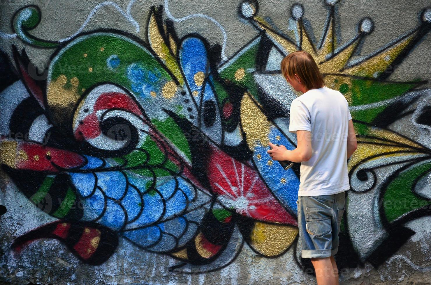 Foto av en ung kille i denim shorts och en vit skjorta. de kille drar på de graffiti vägg en teckning med aerosol målarfärger av olika färger. de begrepp av huliganism och skada till fast egendom