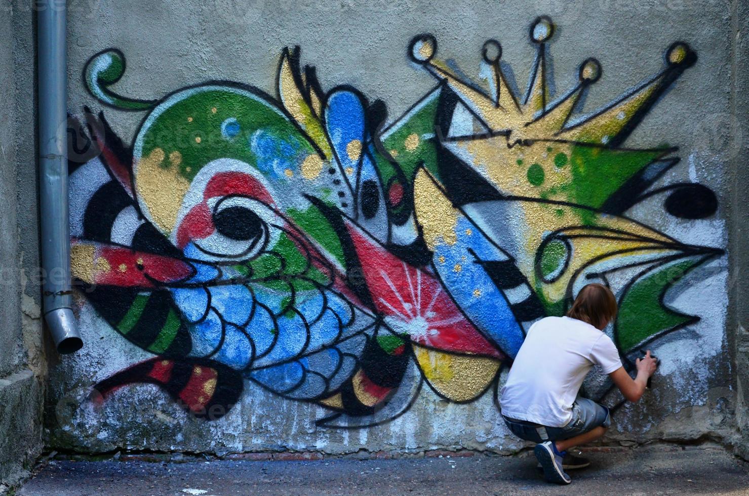 Foto i de bearbeta av teckning en graffiti mönster på ett gammal betong vägg. ung långhårig blond kille drar ett abstrakt teckning av annorlunda färger. gata konst och vandalism begrepp