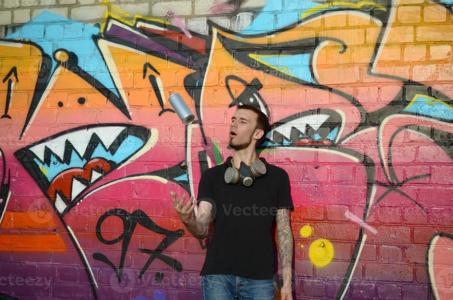 ung graffiti konstnär med gas mask på hans nacke kasta hans spray kan mot färgrik rosa graffiti på tegel vägg. gata konst och samtida målning bearbeta foto