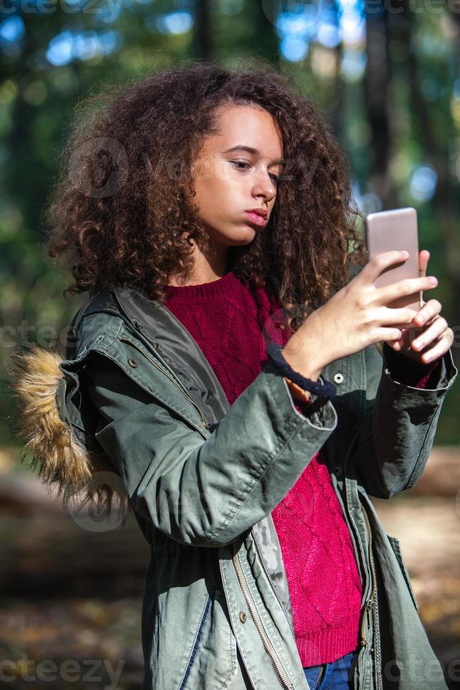söt lockigt hår tonåring flicka med smartphone i höst skog foto