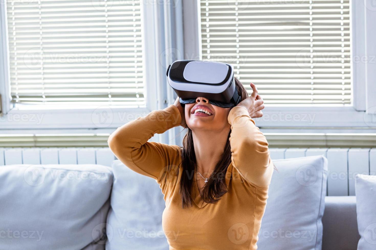 kvinna med virtuell verklighet glasögon. leende ung kvinna använder sig av vr headsetet på Hem på soffa. kvinna njuter virtuell verklighet på Hem. skön kvinna bär vr virtuell verklighet headsetet med gränssnitt. foto