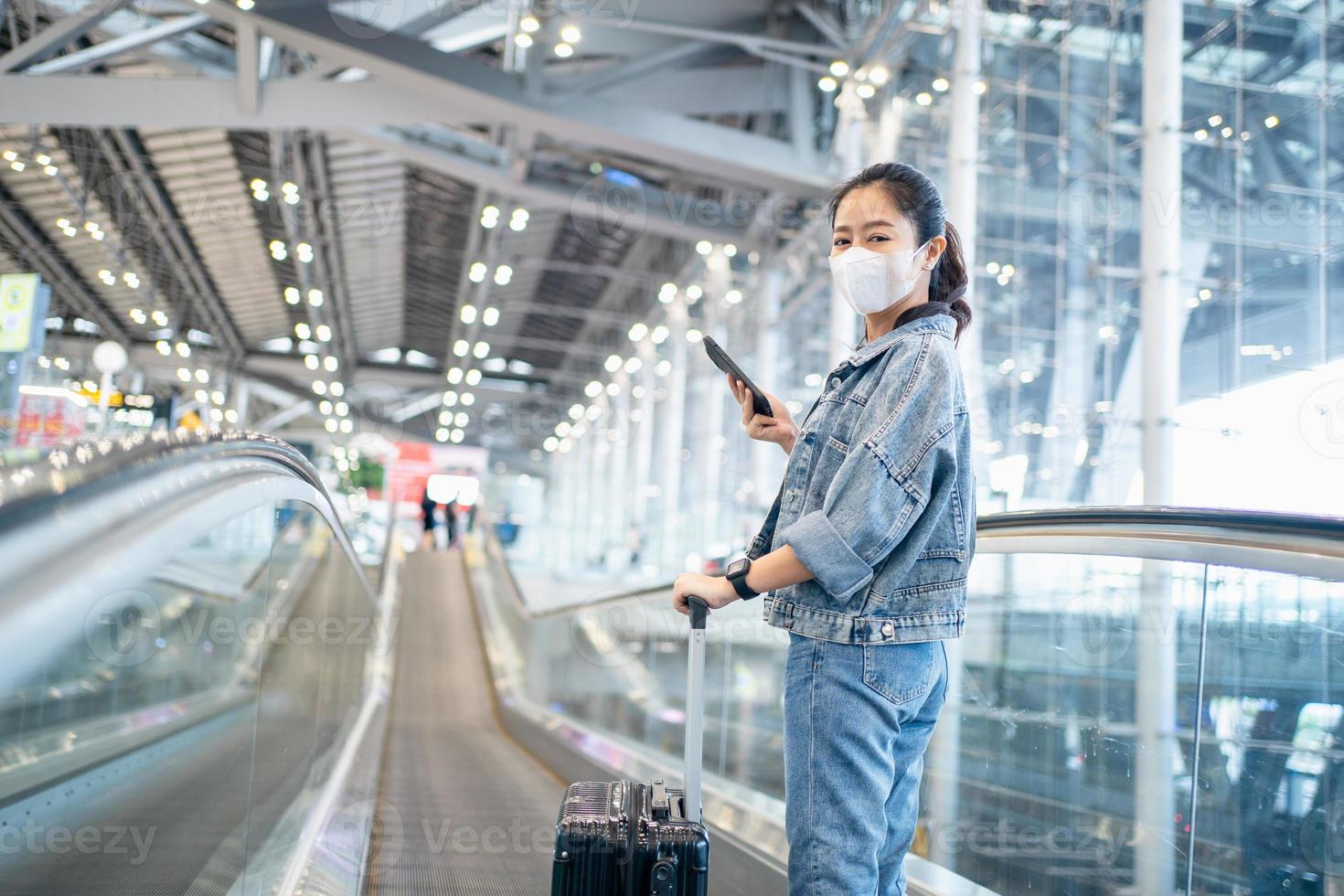 asiatisk kvinna turist bär ansikte mask och innehav smartphone på rulltrappa på flygplats terminal under coronavirus eller covid-19 utbrott. foto