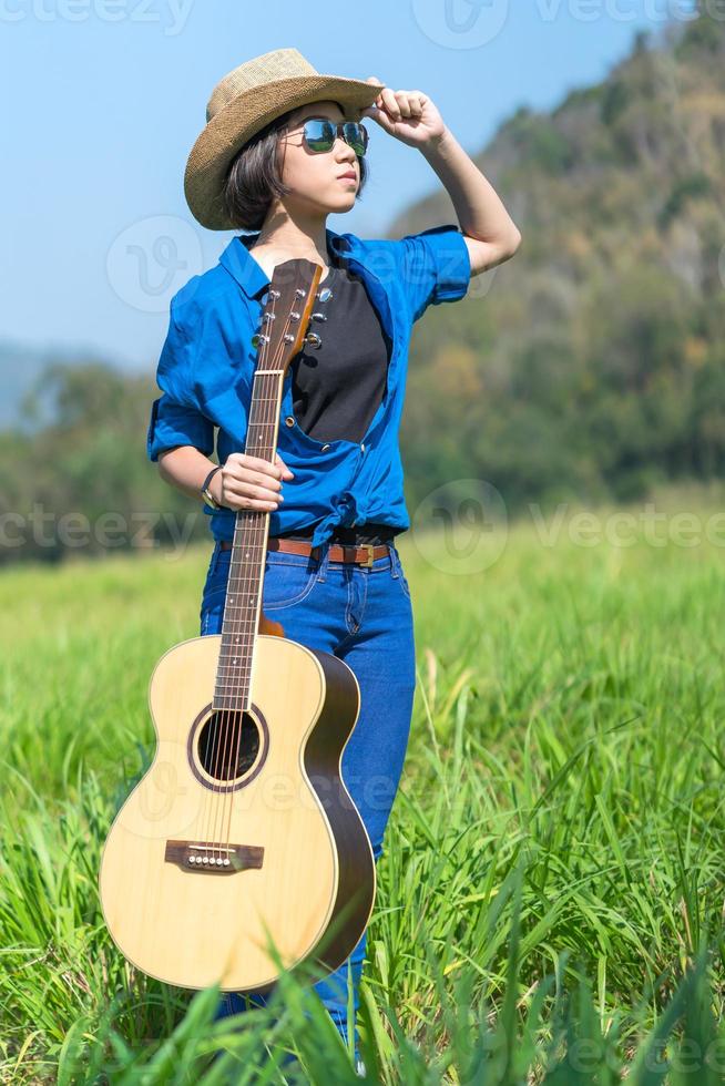 kvinna ha på sig hatt och bära henne gitarr i gräs fält foto