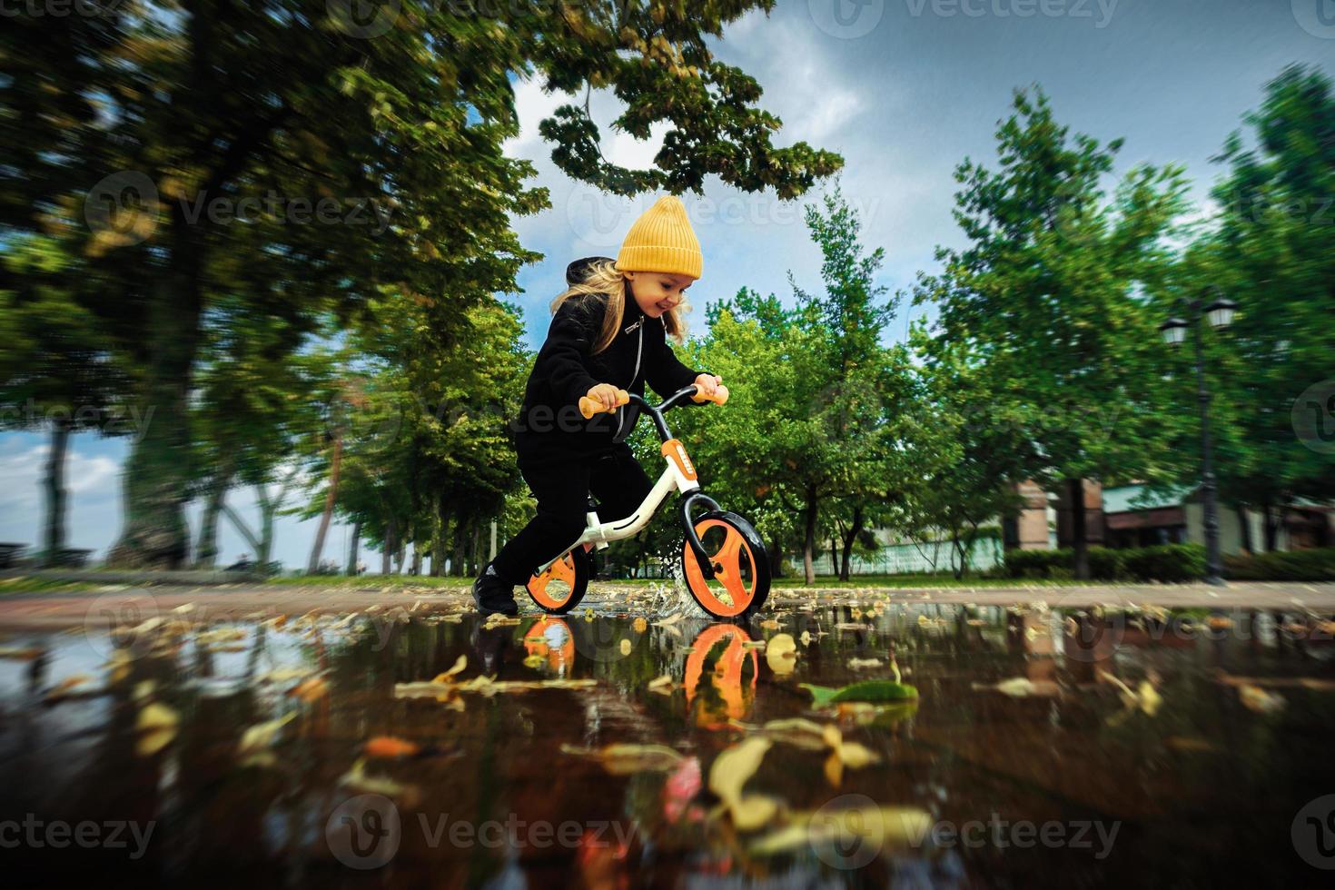 sötnos liten flicka rider genom de vattenpölar på cykel på de parkera foto