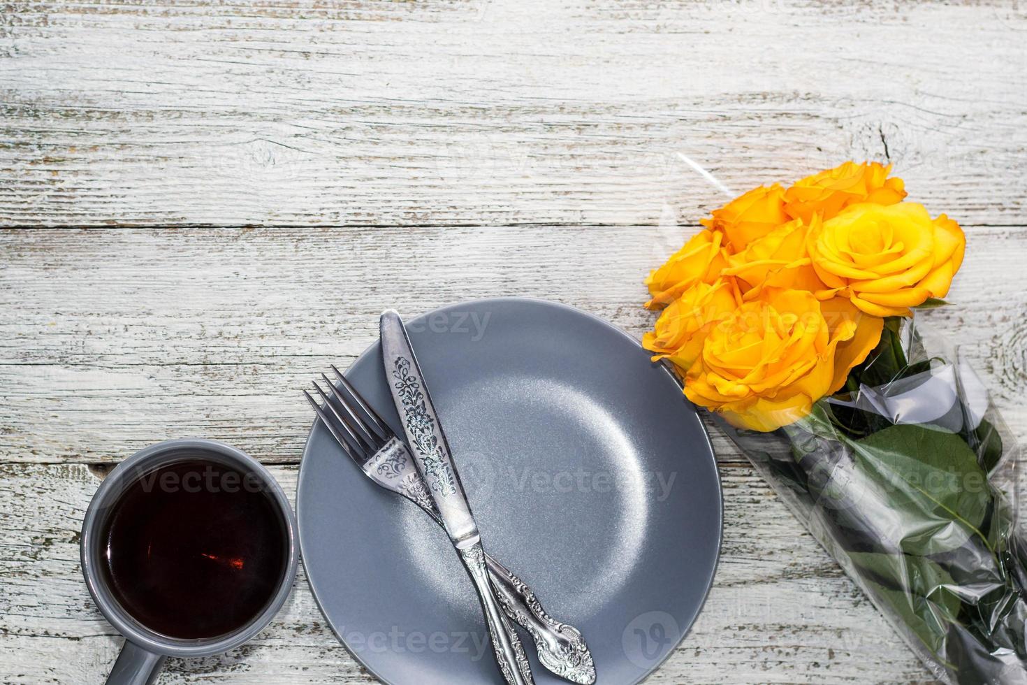 en tallrik med en kniv och gaffel och te med en bukett av gul ro på en vit trä- bakgrund foto