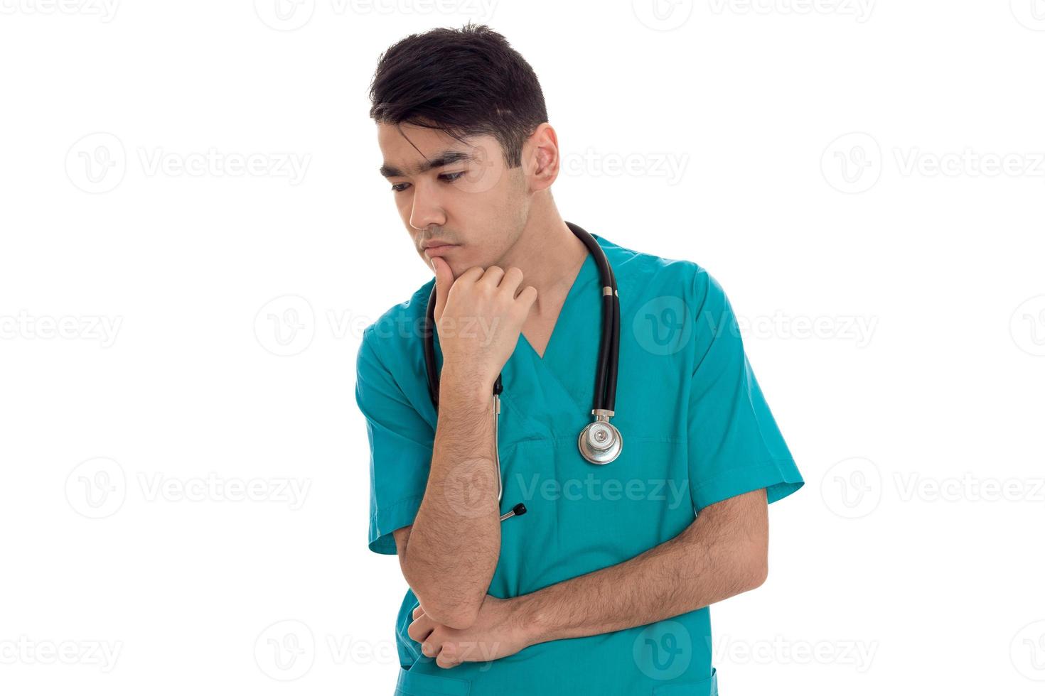 ung omtänksam brunett man läkare i blå enhetlig med stetoskop på hans axlar tänkande isolerat på vit bakgrund foto