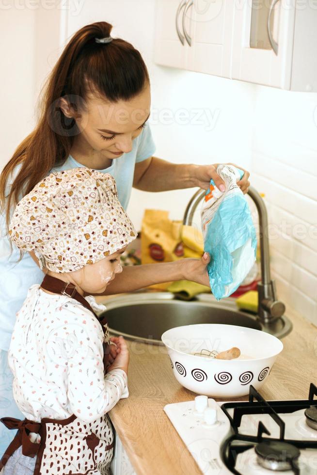 söt liten hemkock flicka med henne skön mor gör pannkakor i vit kök. foto