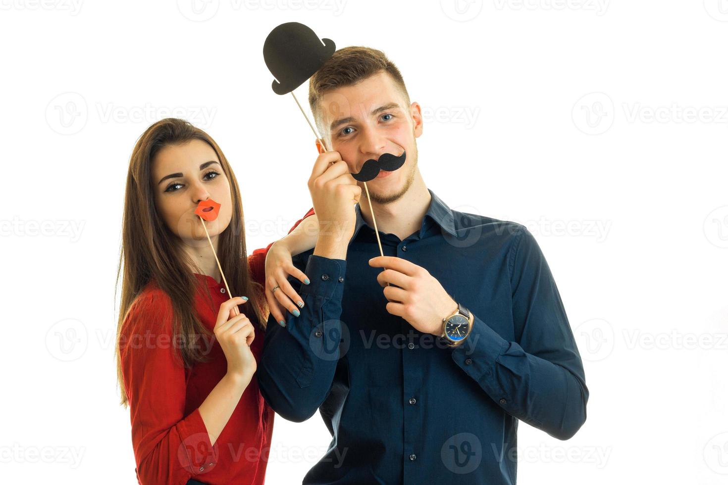 ung skön flicka och kille håll nära ansikte papper rekvisita i de form av en mustasch, mun och hatt och Framställ för de kamera foto