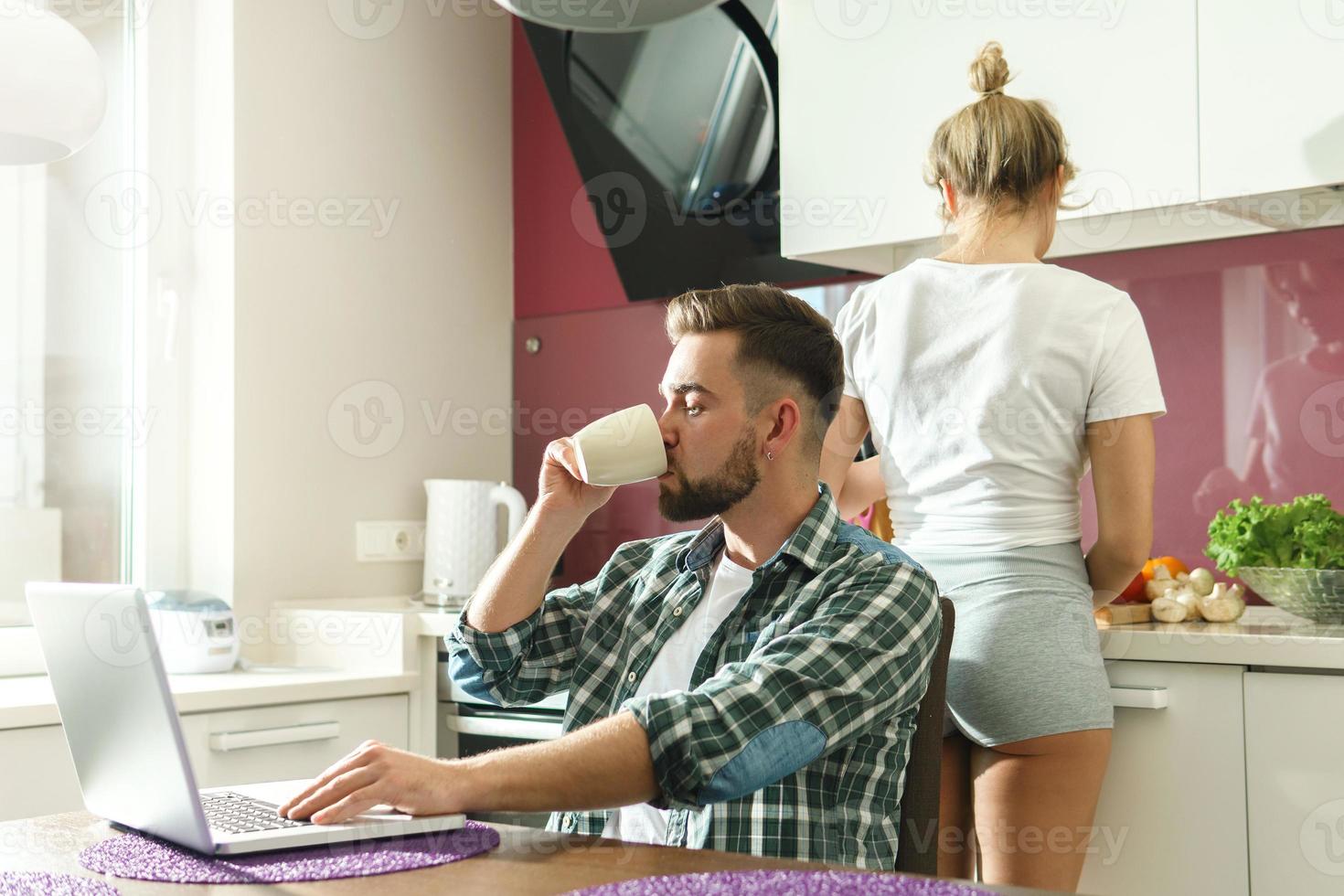 Lycklig par i de kök på morgon. stilig man använder sig av bärbar dator medan hans fru matlagning frukost. foto