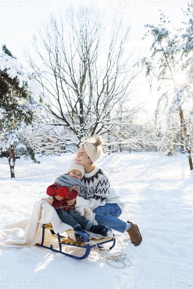 ung mor och henne söt liten son med retro kälke i en snöig parkera under solig dag foto