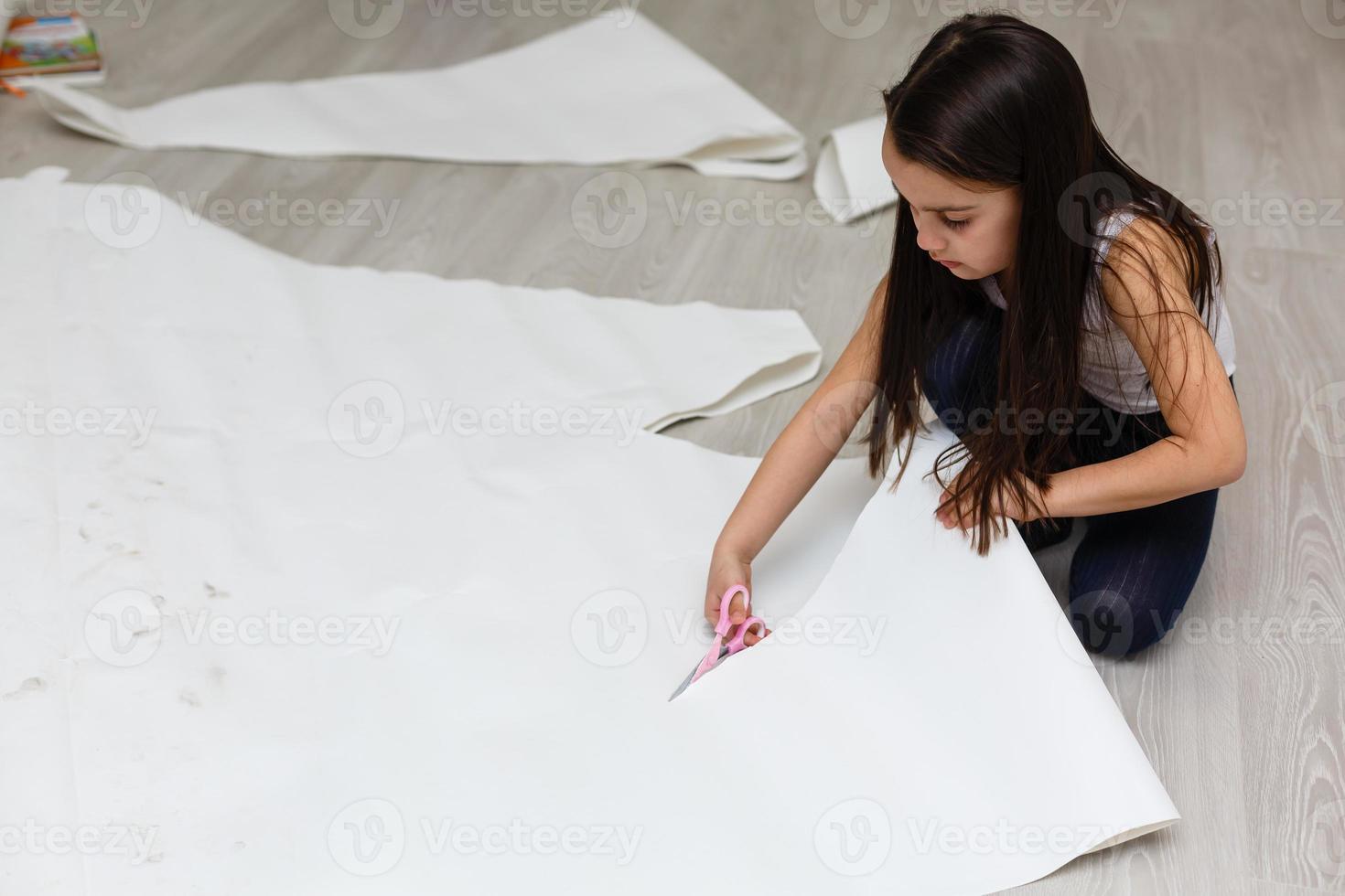 liten flicka är skärande papper använder sig av sax medan Sammanträde foto