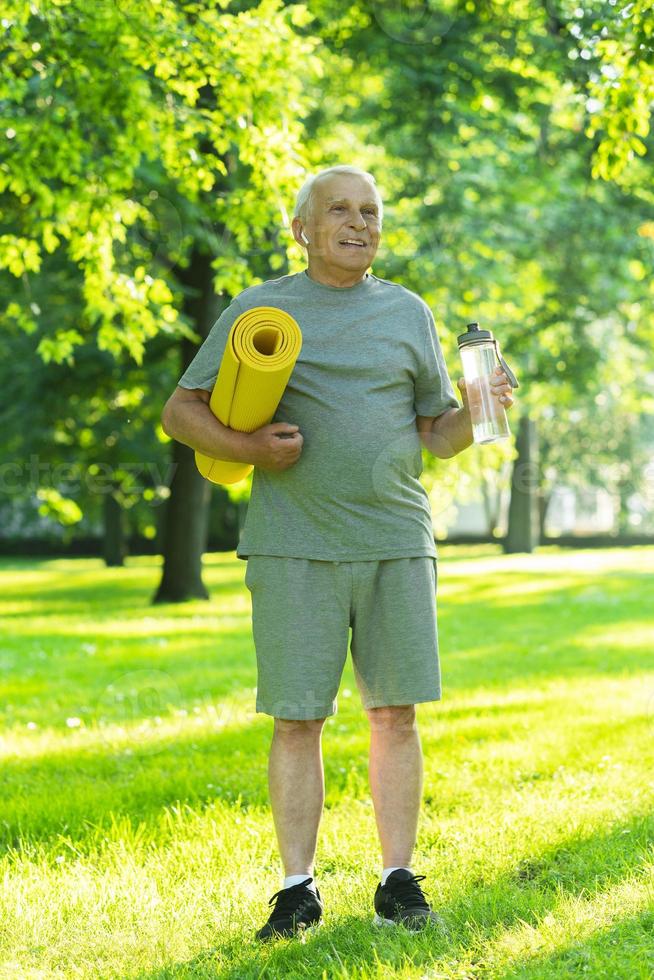 aktiva äldre man med en övning matta och flaska av vatten i grön stad parkera foto