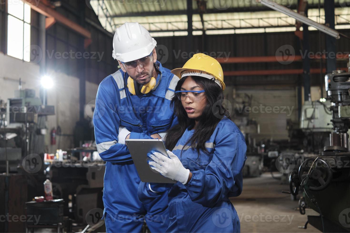 två industriell arbetare i säkerhet uniformer och hårda hattar, manlig chef, och svart kollega arbete med läsplatta till kolla upp metallarbete maskiner i tillverkning fabrik. professionell produktion ingenjör team. foto